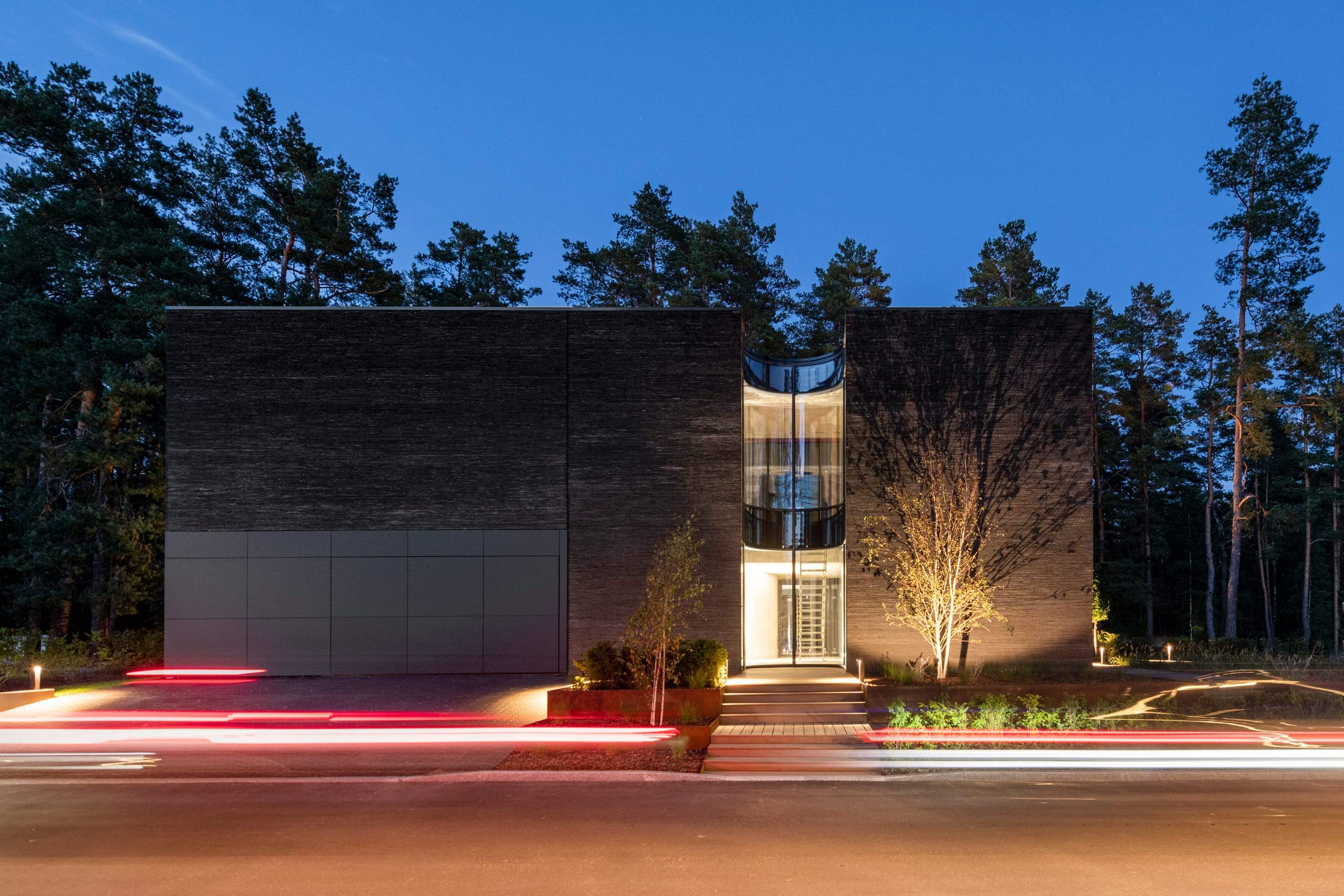 IN JAN House — lit at night, dark stone volumes behind a road streaked with car light trails
