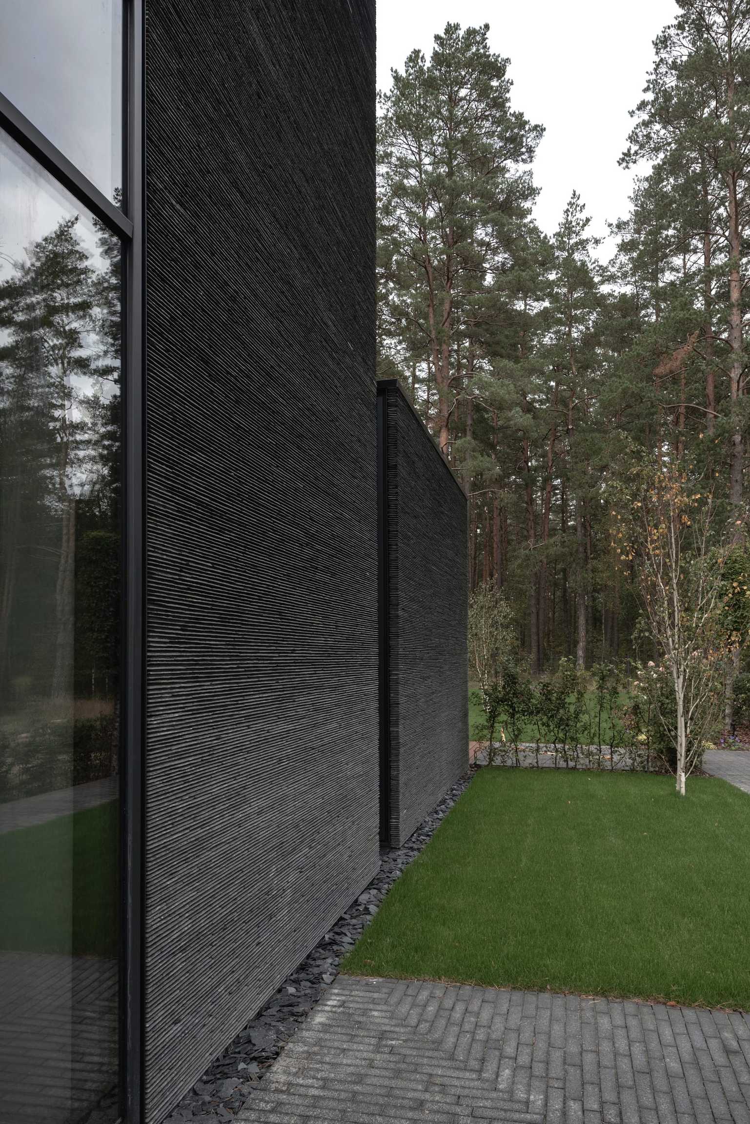 IN JAN House — ribbed dark stone cladding running from a glazed corner towards the pines