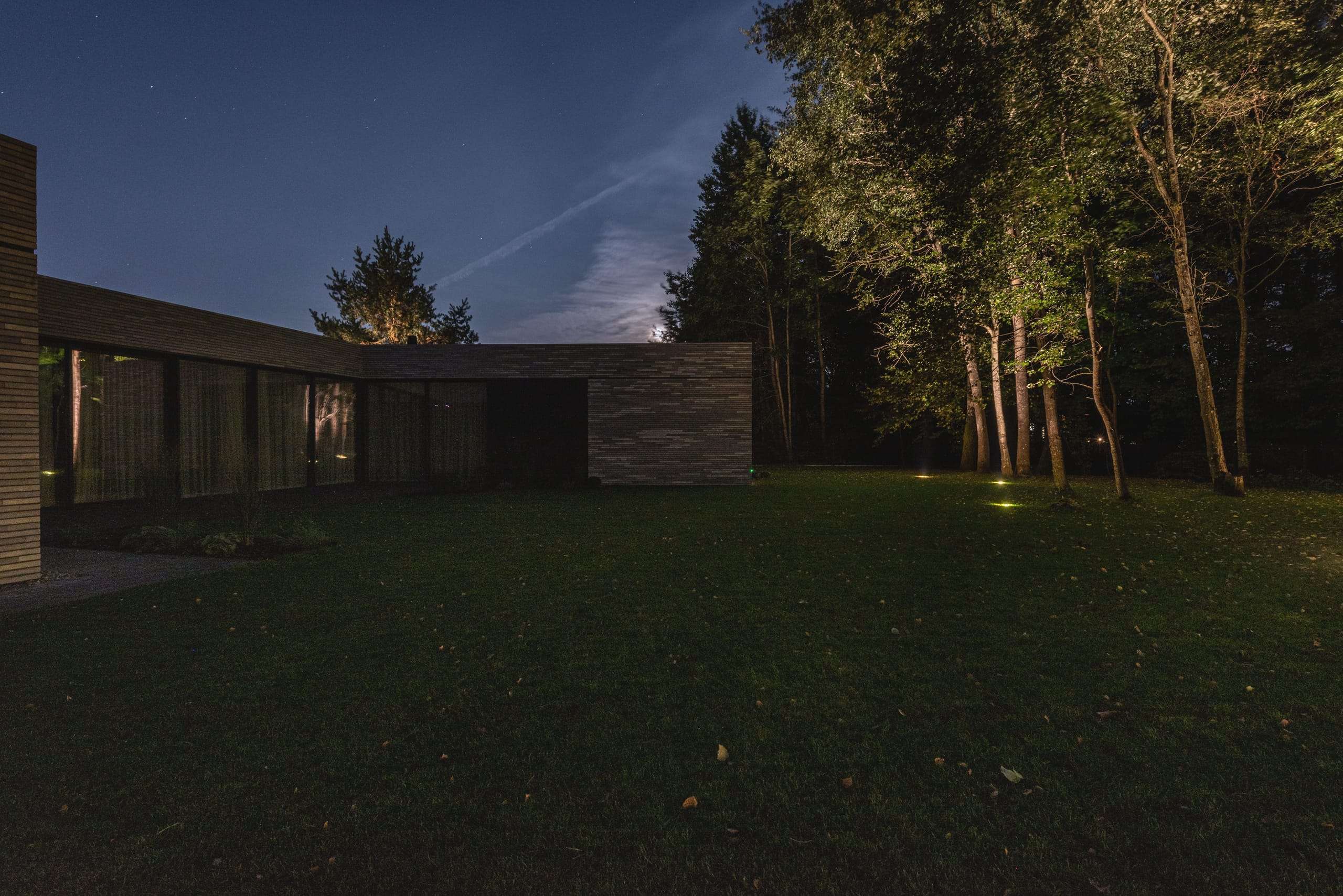 Modern Family House — at night seen across the lawn, uplit trees along the boundary