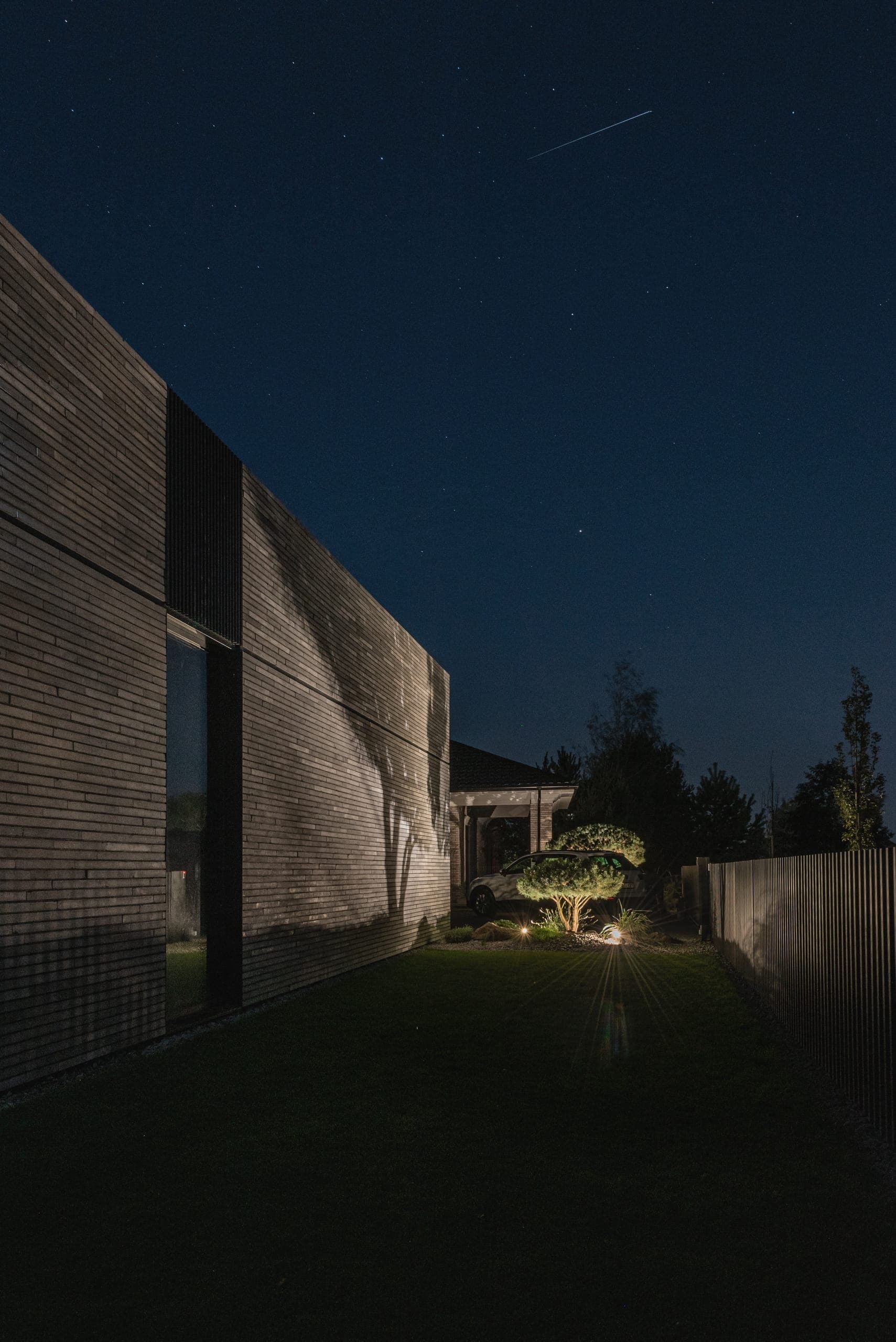 Modern Family House — long brick wall at night, a lit topiary pine at the garden's end